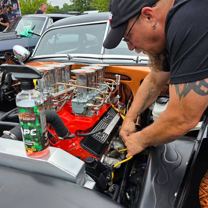 A man in a cap leans over an open car hood with a bottle of APC to his left