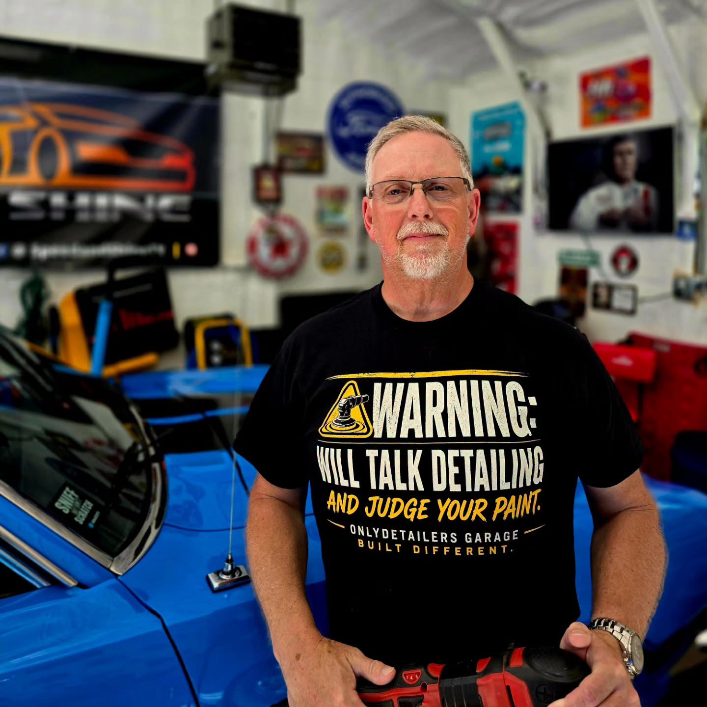 A man wearing a black graphic t-shirt stands in a garage in front of a bright blue car. The shirt features a yellow and white warning graphic that reads 'WARNING: WILL TALK DETAILING AND JUDGE YOUR PAINT.