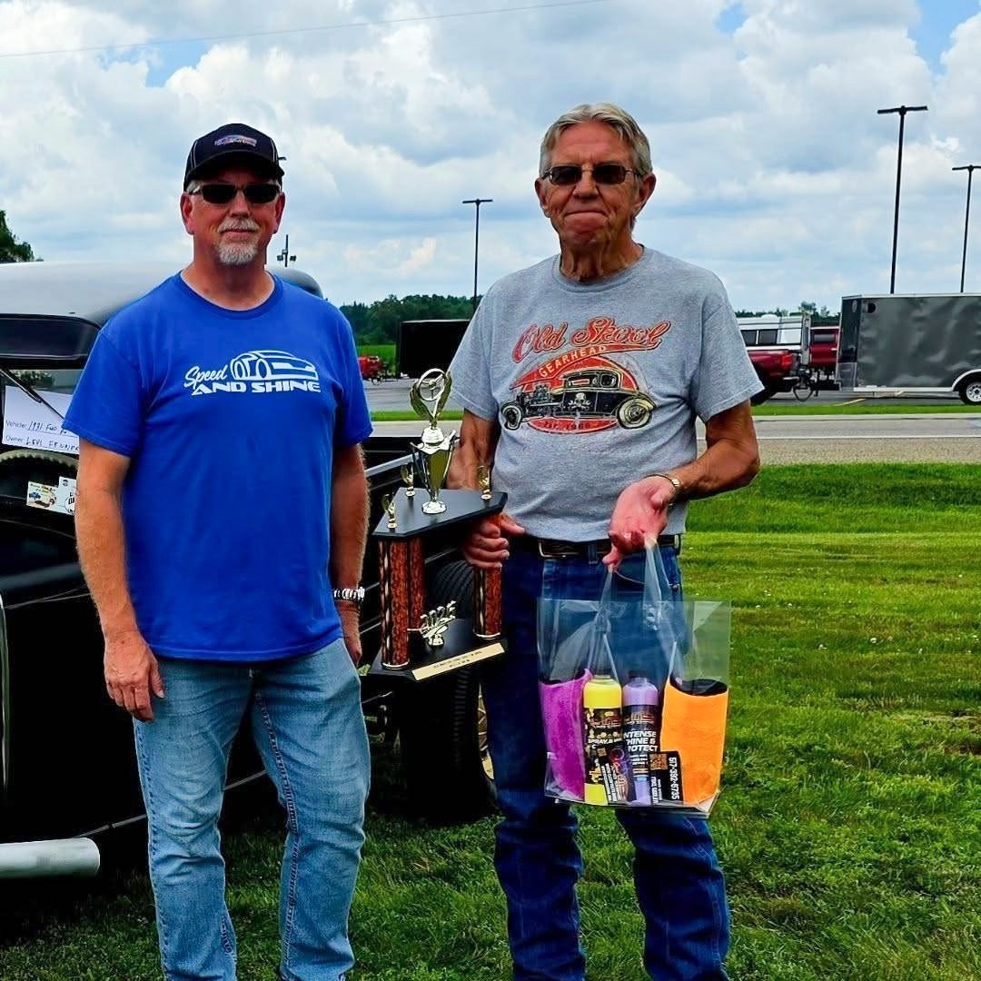 Two men standing outdoors on a grassy field holding a trophy and a Speed And Shine gift bag.